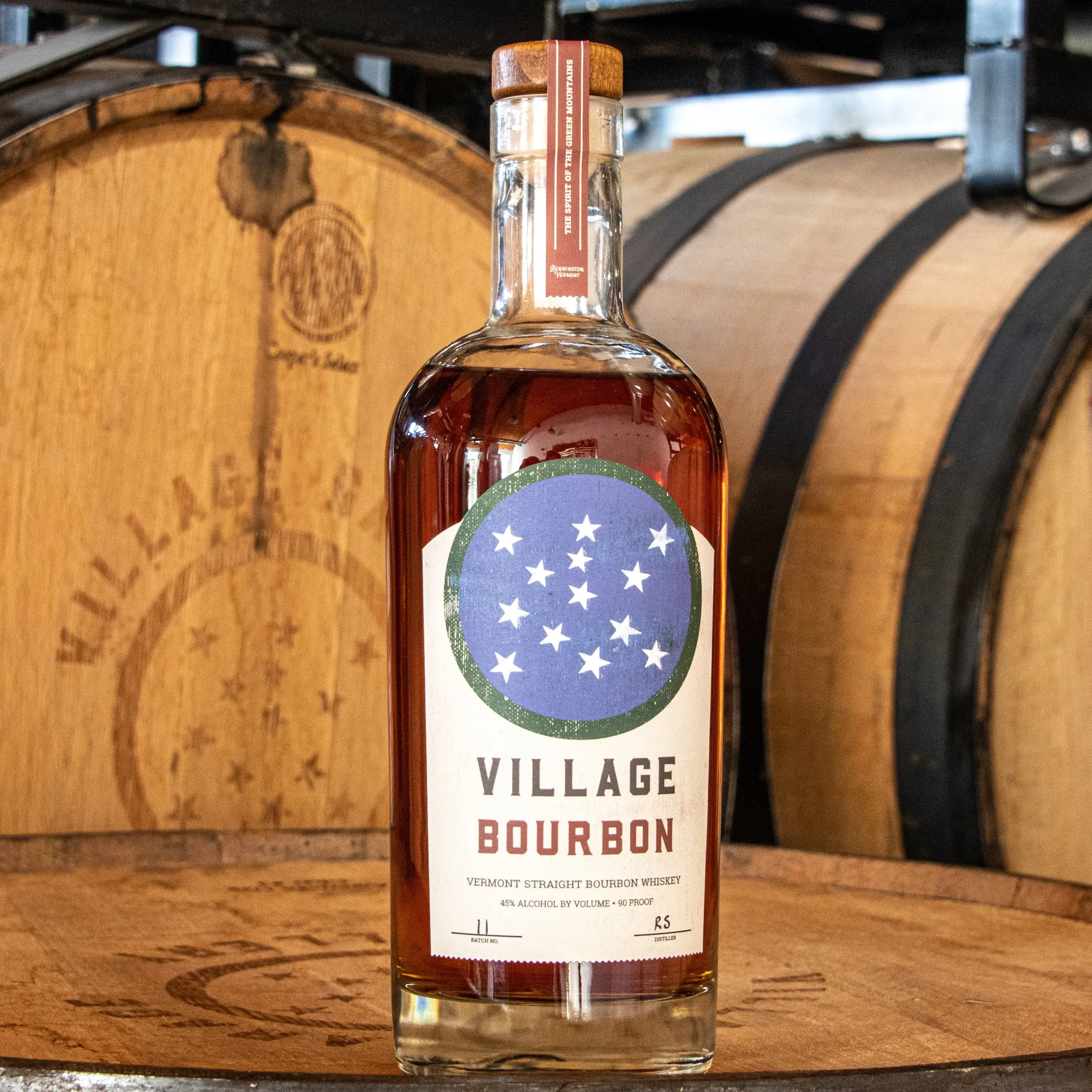 Bottle of Village Bourbon straight bourbon whiskey in front of oak barrels